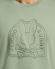 The MENS MELBOURNE GRAPHIC TEE - B6U410F200 by Psycho Bunny is a sage green shirt made from eco-friendly Peruvian Pima cotton, showcasing an embossed bunny head and crossed bones design encircled by "Psycho Bunny.