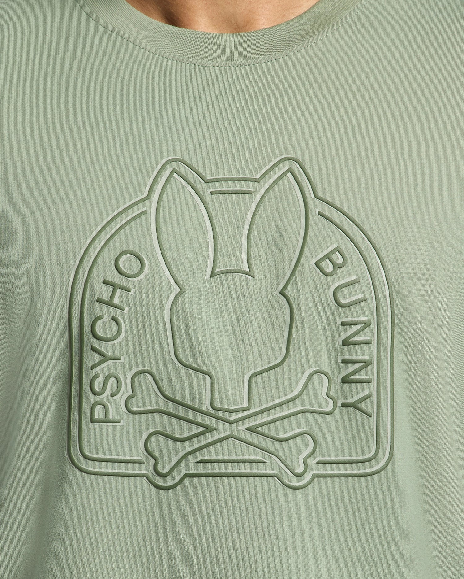 The MENS MELBOURNE GRAPHIC TEE - B6U410F200 by Psycho Bunny is a sage green shirt made from eco-friendly Peruvian Pima cotton, showcasing an embossed bunny head and crossed bones design encircled by "Psycho Bunny.