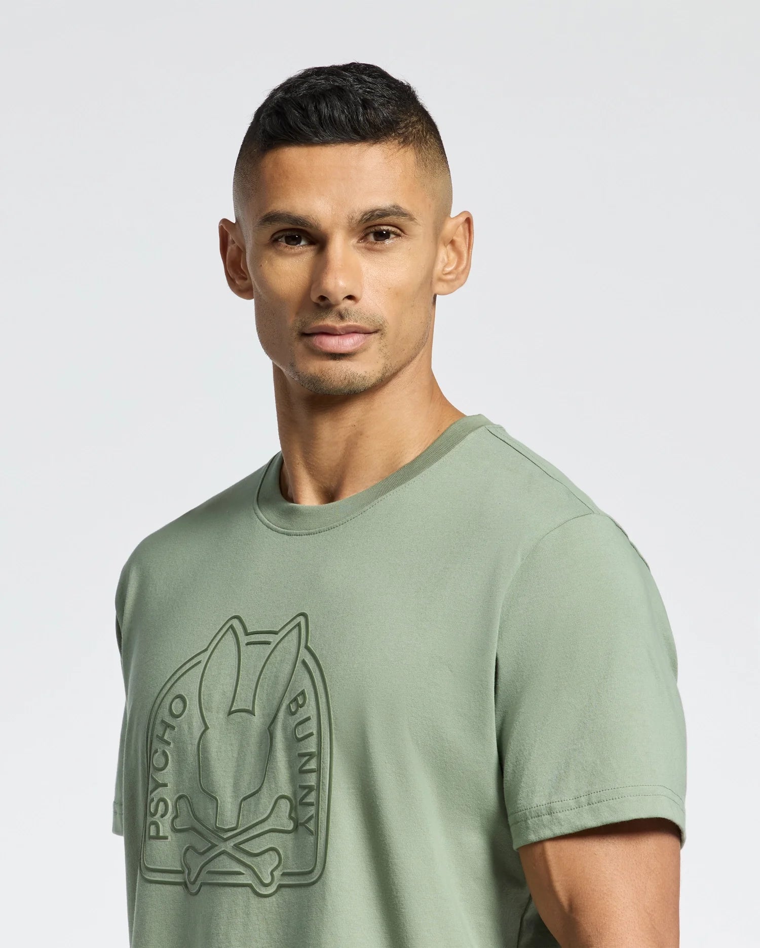 A person with short dark hair and a neutral expression wears the MENS MELBOURNE GRAPHIC TEE - B6U410F200 by Psycho Bunny. Made from eco-friendly Peruvian Pima cotton, it features a raised logo on the front. The white background accentuates the green tee.