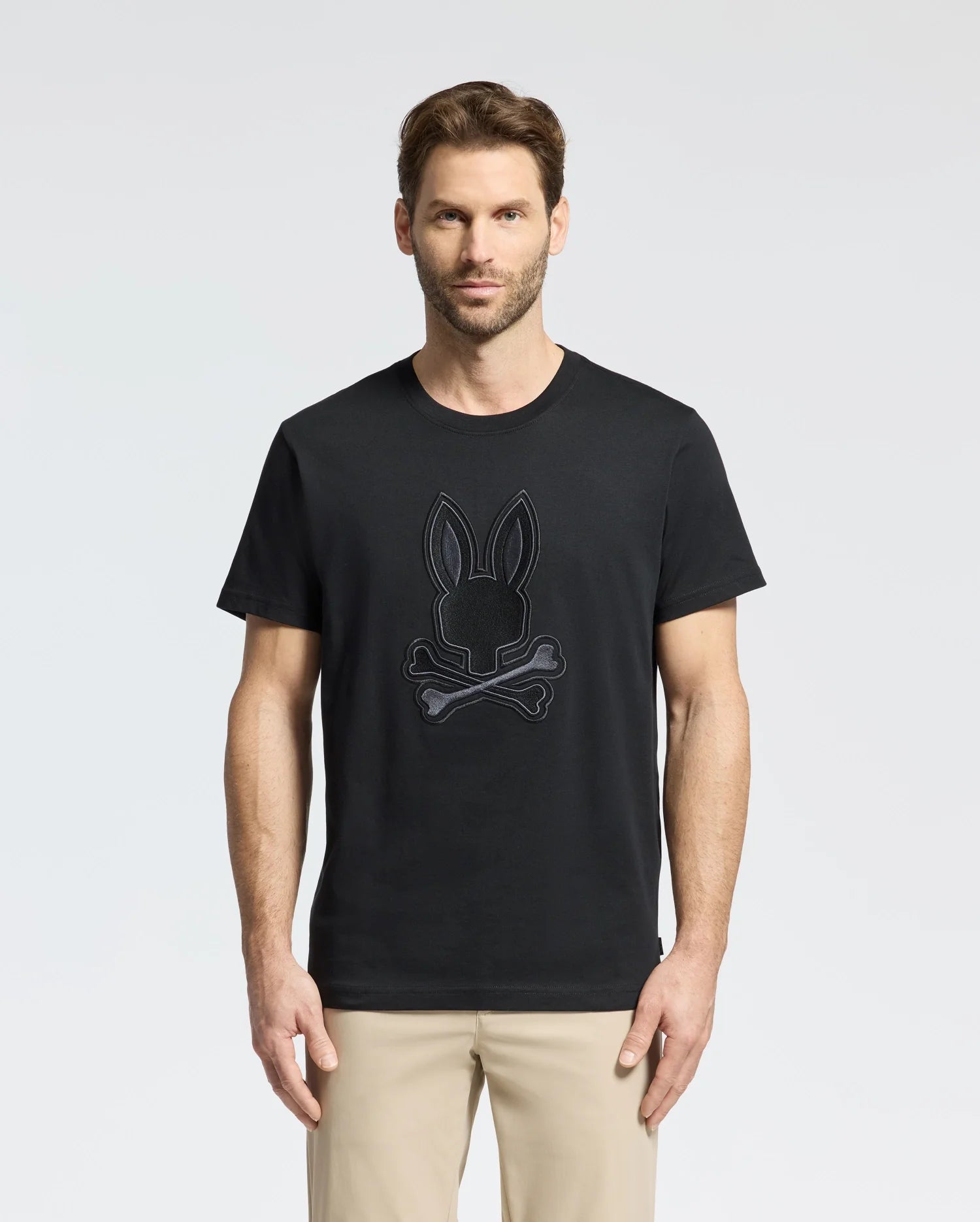 A man with short brown hair and a beard wears the Psycho Bunny MENS NEW CASTLE GRAPHIC TEE - B6U415F200, made from soft Pima cotton with an embroidered bunny ears and crossbones design. He pairs it with beige pants, standing against a plain white background.