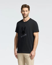 A man with short brown hair and a beard wears the black Psycho Bunny MENS NEW CASTLE GRAPHIC TEE featuring an embroidered bunny and crossbones. Against a plain white background, he pairs it casually with beige pants.
