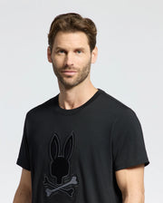 A man with short brown hair models the Psycho Bunny MENS NEW CASTLE GRAPHIC TEE - B6U415F200, adorned with a bunny and crossbones design, against a plain background.