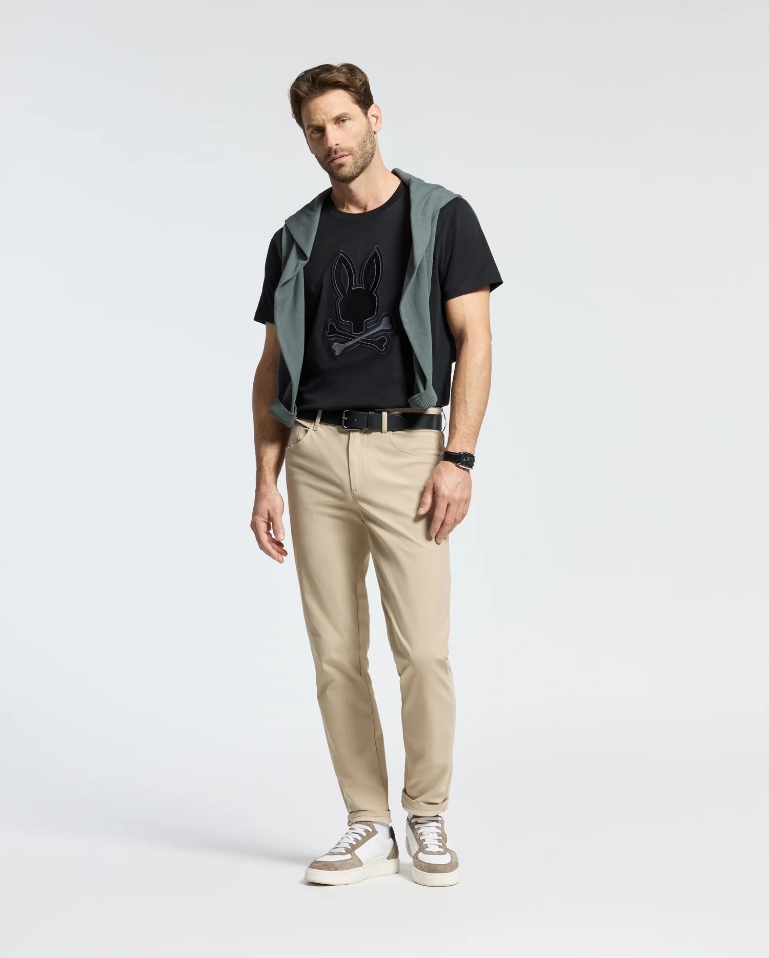 A man stands against a plain background in Psycho Bunny's Men's New Castle Graphic Tee - B6U415F200 made from soft Pima cotton, paired with beige pants and white sneakers. A dark jacket drapes over his shoulders; with one hand in his pocket, he exudes effortless style.