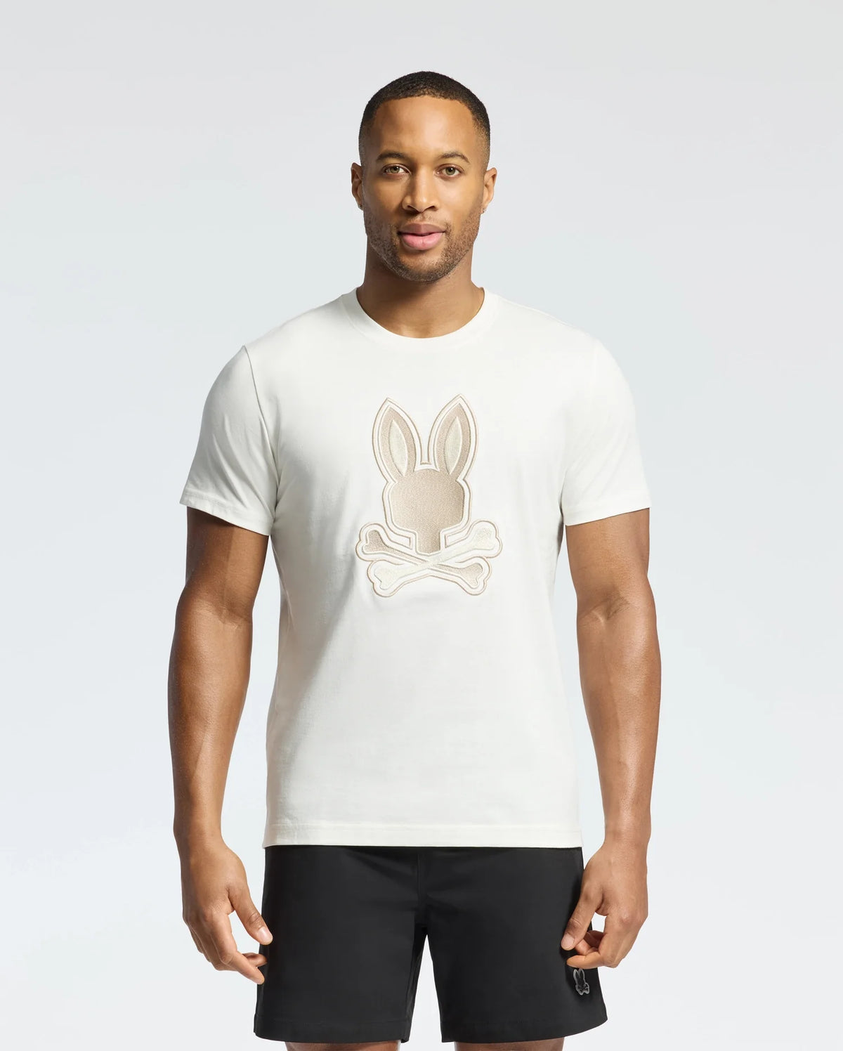 A man wears a Psycho Bunny New Castle graphic tee, B6U415F200, made from soft Pima cotton with an embroidered bunny and crossed bones. He pairs it with black shorts against a plain, light background.
