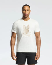 A man wears a Psycho Bunny New Castle graphic tee, B6U415F200, made from soft Pima cotton with an embroidered bunny and crossed bones. He pairs it with black shorts against a plain, light background.