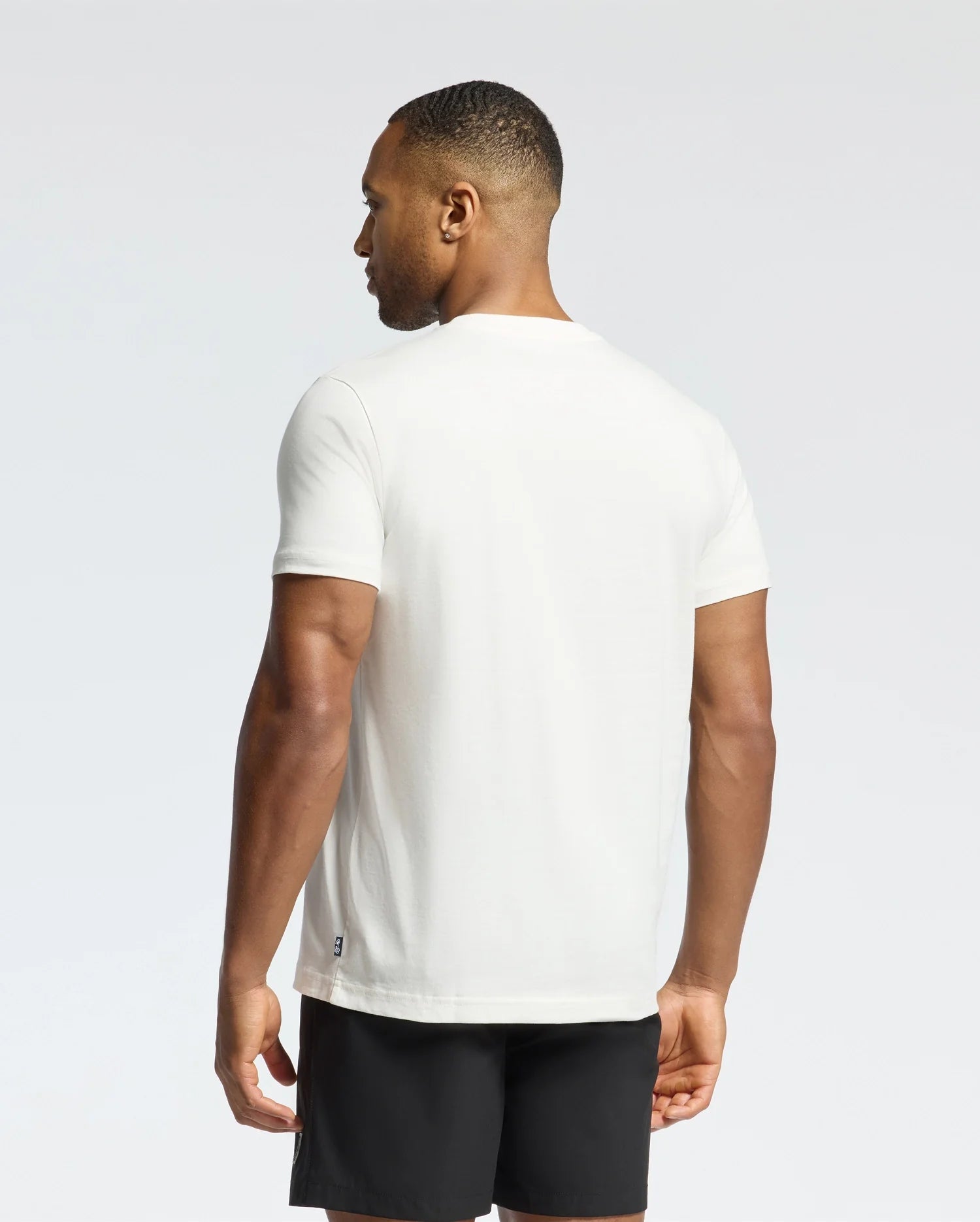 A man wearing a Psycho Bunny MENS NEW CASTLE GRAPHIC TEE and black shorts stands sideways to the camera, his head turned slightly left against a plain white background.