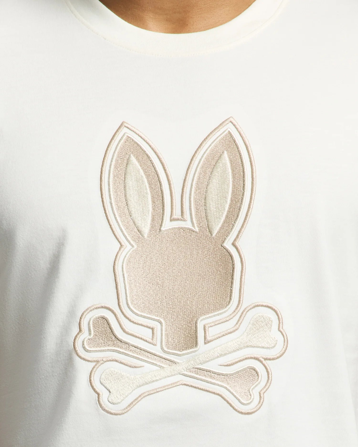 The MENS NEW CASTLE GRAPHIC TEE by Psycho Bunny is a white Pima cotton T-shirt featuring a beige embroidered bunny logo with a stylized head above two crossed bones, offering a playful twist on the classic skull and crossbones design.