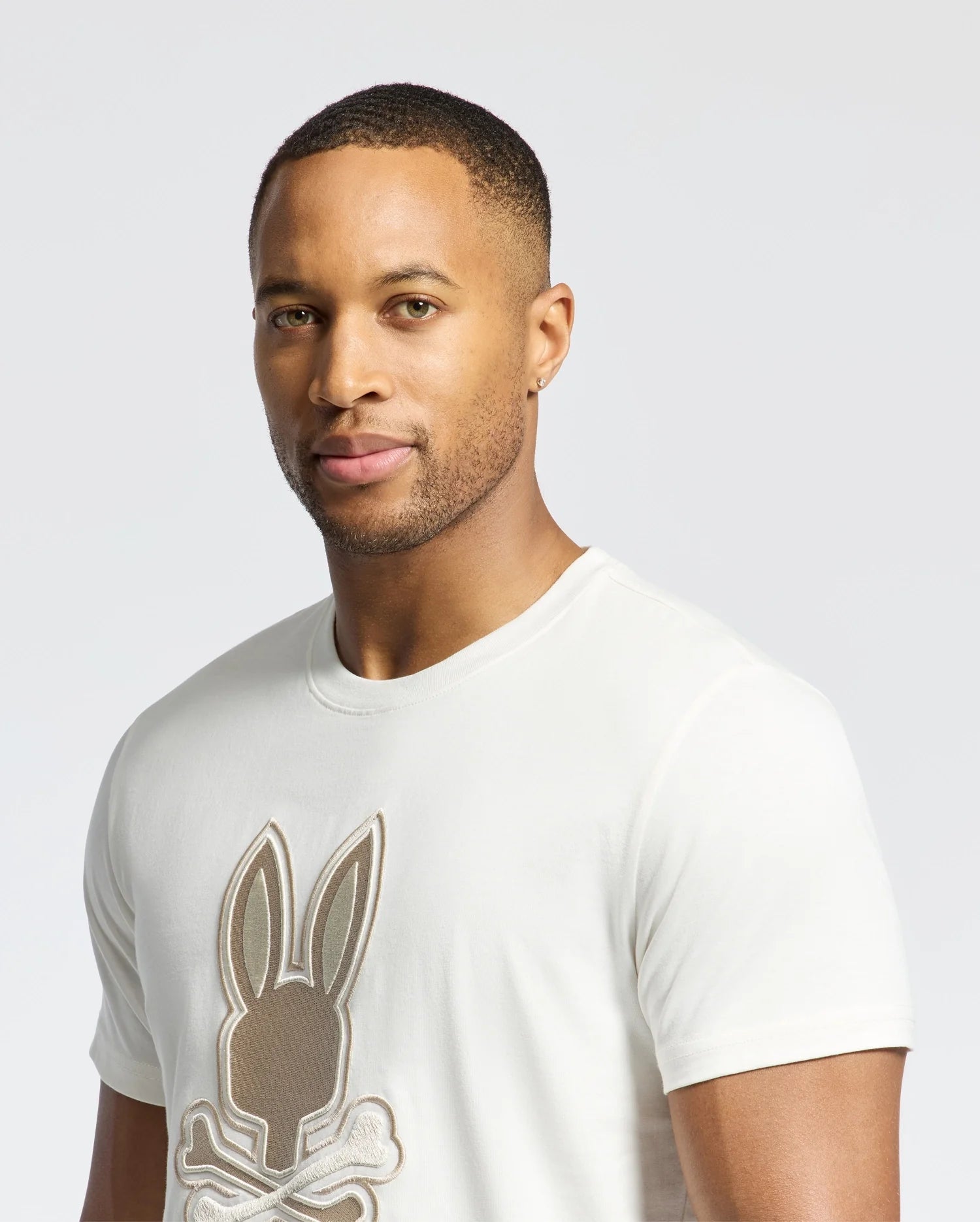 A man stands against a plain backdrop wearing a light-colored Psycho Bunny MENS NEW CASTLE GRAPHIC TEE (B6U415F200) with an embroidered bunny logo.