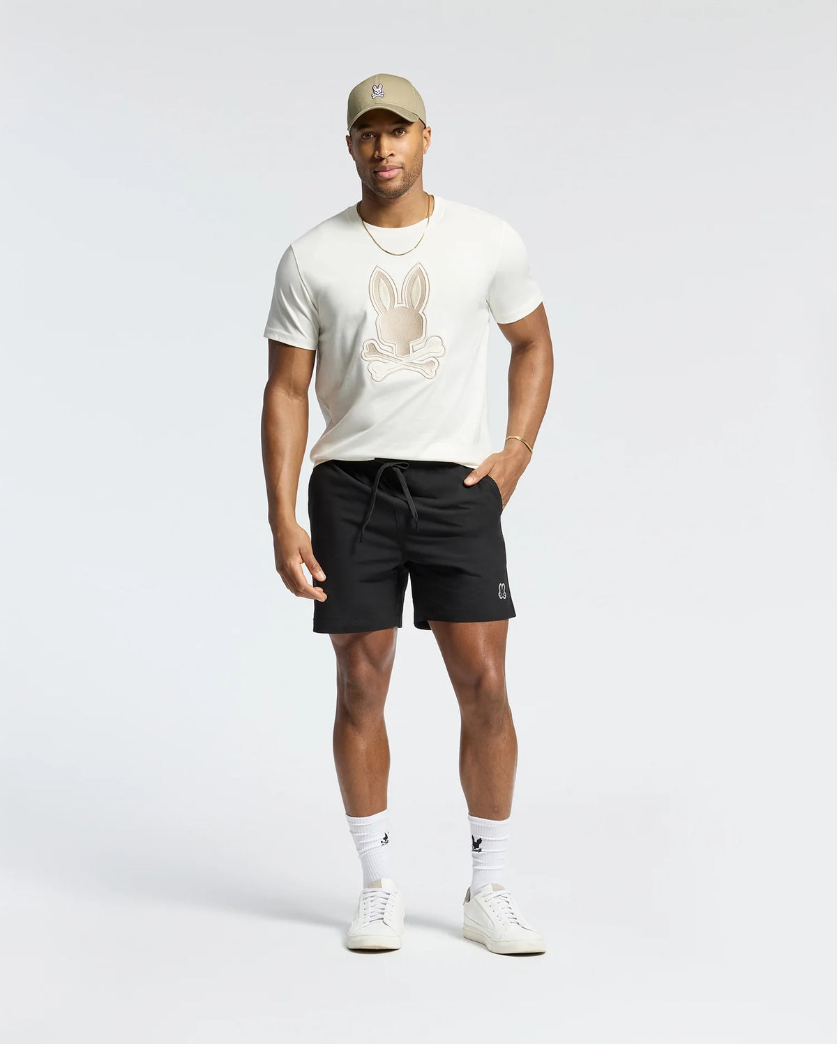 A man wearing a beige cap, a Psycho Bunny Pima cotton tee (MENS NEW CASTLE GRAPHIC TEE - B6U415F200) with an embroidered bunny logo, black shorts, white socks, and white sneakers stands against a plain background.