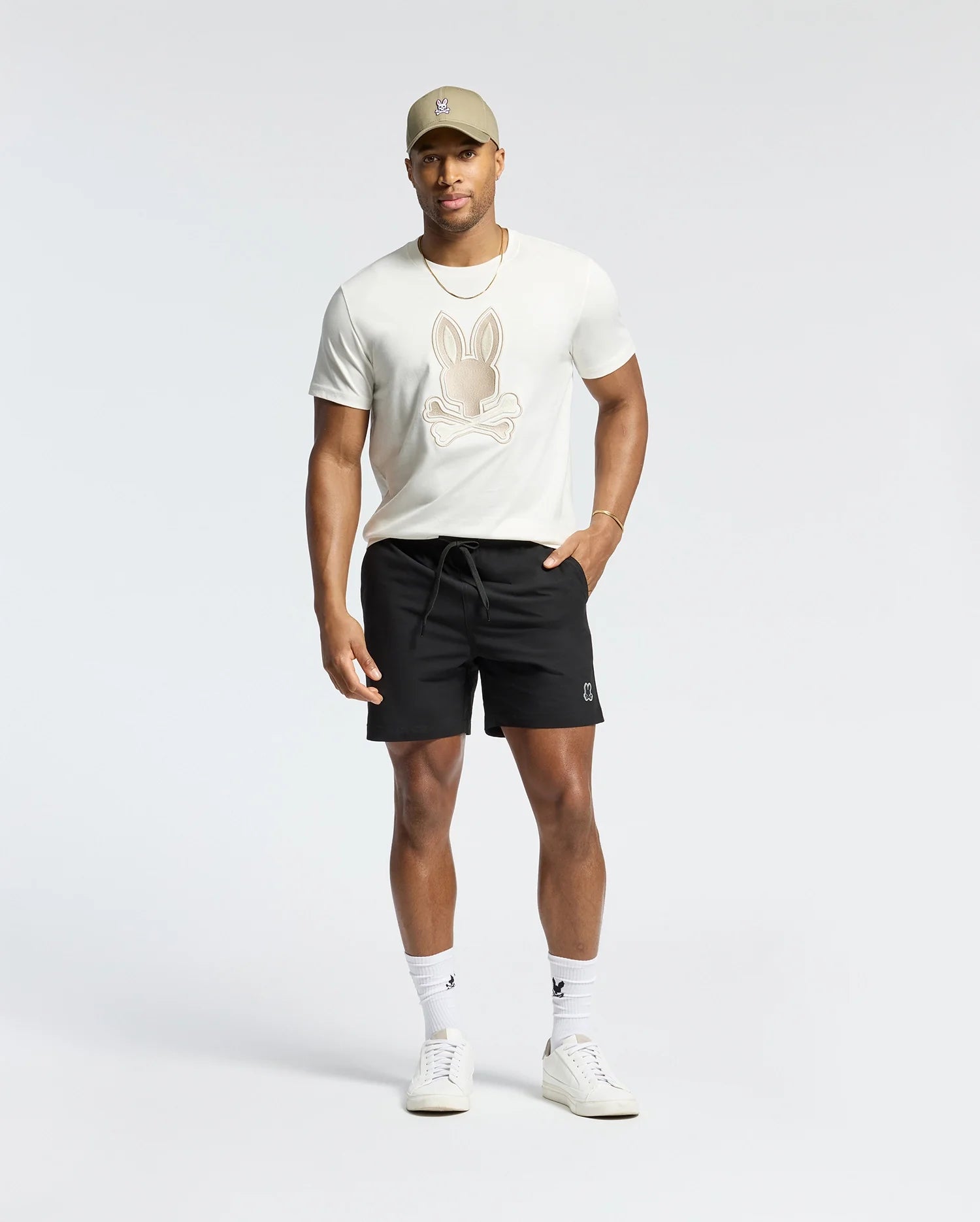 A man wearing a beige cap, a Psycho Bunny Pima cotton tee (MENS NEW CASTLE GRAPHIC TEE - B6U415F200) with an embroidered bunny logo, black shorts, white socks, and white sneakers stands against a plain background.
