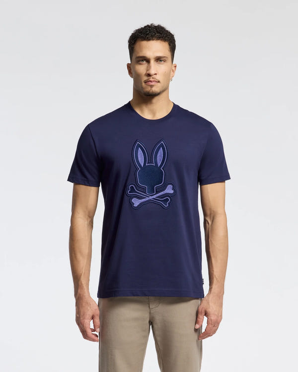 A man in a Psycho Bunny MENS NEW CASTLE GRAPHIC TEE (B6U415F200) with a bunny and crossbones design stands against a neutral background. The soft Pima cotton tee pairs perfectly with light khaki pants. His short curly hair enhances the effortlessly casual look.