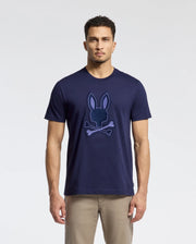A man in a Psycho Bunny MENS NEW CASTLE GRAPHIC TEE (B6U415F200) with a bunny and crossbones design stands against a neutral background. The soft Pima cotton tee pairs perfectly with light khaki pants. His short curly hair enhances the effortlessly casual look.