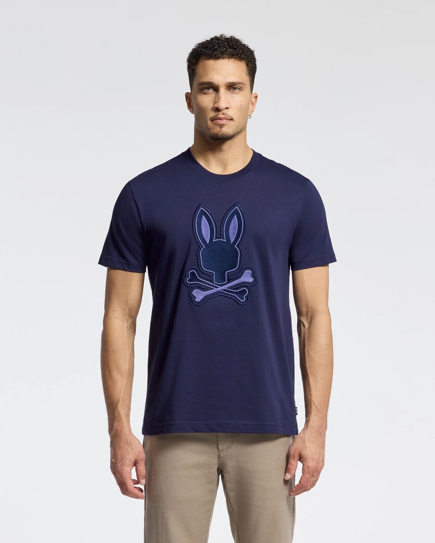 A man in a Psycho Bunny MENS NEW CASTLE GRAPHIC TEE (B6U415F200) with a bunny and crossbones design stands against a neutral background. The soft Pima cotton tee pairs perfectly with light khaki pants. His short curly hair enhances the effortlessly casual look.
