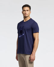 A man models Psycho Bunny's MENS NEW CASTLE GRAPHIC TEE (B6U415F200), made of navy blue Pima cotton with an embroidered bunny skull and crossbones design, against a white backdrop. He sports short curly hair, a slight beard, and pairs the tee with light khaki pants.