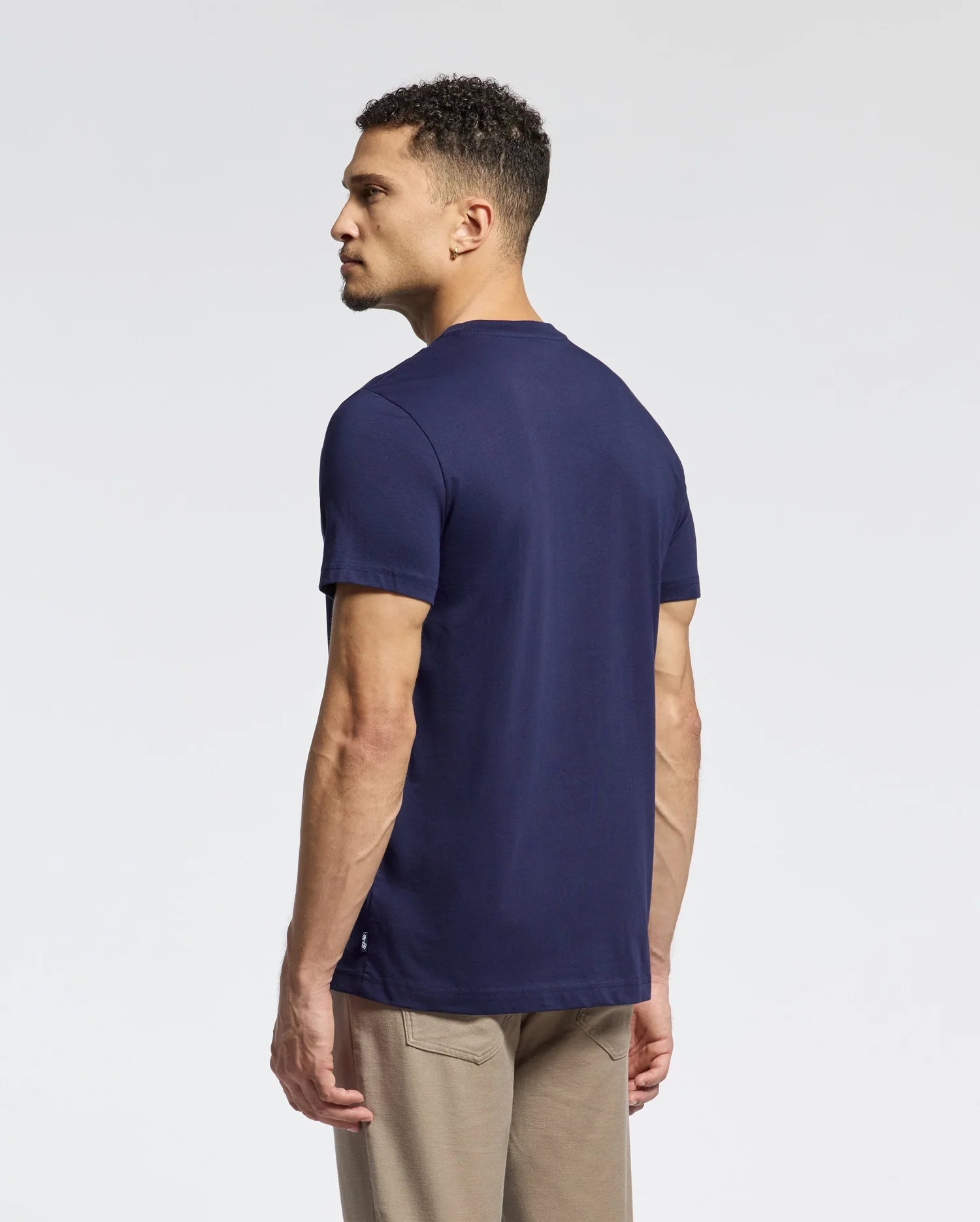 A man in a Psycho Bunny MENS NEW CASTLE GRAPHIC TEE (B6U415F200) and beige pants stands sideways, gazing left against a light gray background.