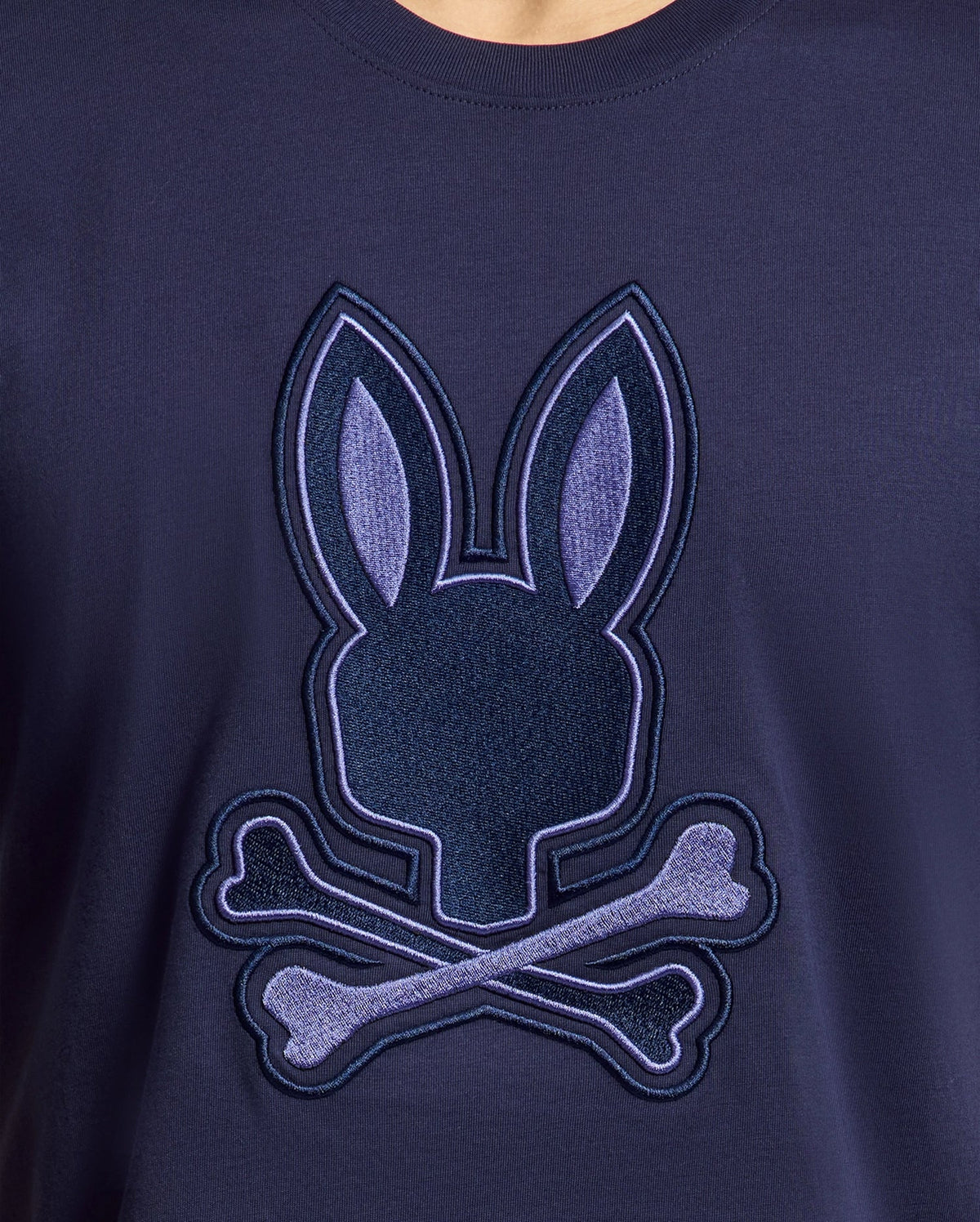 Close-up of the Psycho Bunny MENS NEW CASTLE GRAPHIC TEE - B6U415F200 in navy blue, made from soft Pima cotton. It features an embroidered bunny head with crossed bones, resembling a skull motif in shades of blue with stitched details.