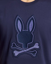 Close-up of the Psycho Bunny MENS NEW CASTLE GRAPHIC TEE - B6U415F200 in navy blue, made from soft Pima cotton. It features an embroidered bunny head with crossed bones, resembling a skull motif in shades of blue with stitched details.