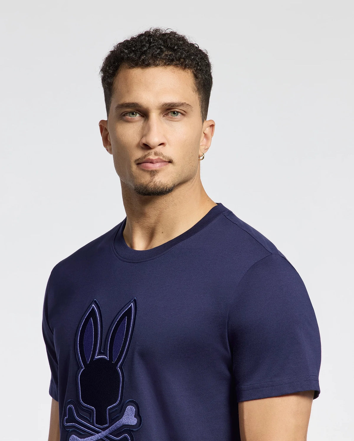 A man with short curly hair and a goatee wears Psycho Bunny's MENS NEW CASTLE GRAPHIC TEE, featuring a rabbit and crossbones logo. Made from soft Pima cotton, the navy blue shirt complements his left ear earring as he poses against a plain background.