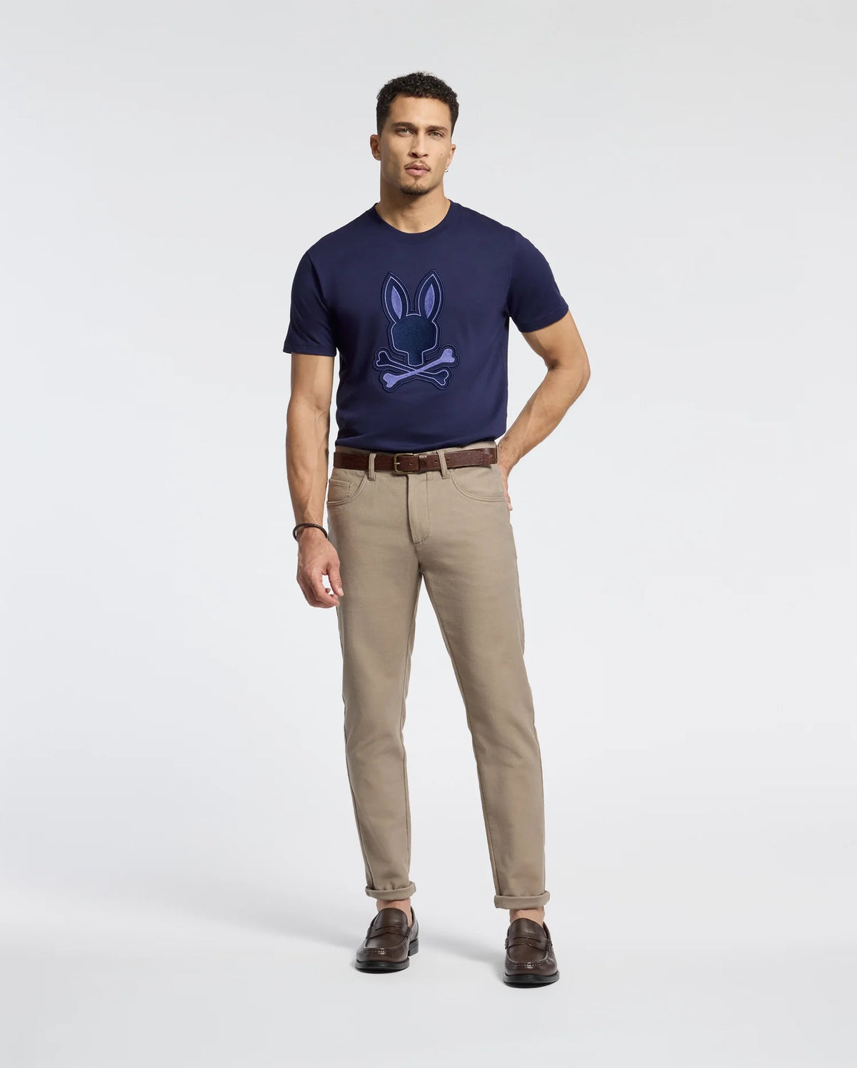 A man wears the Psycho Bunny MENS NEW CASTLE GRAPHIC TEE - B6U415F200, a navy blue Pima cotton tee with an embroidered bunny design, against a white background. He completes his look with beige pants, brown leather shoes, and a matching belt.