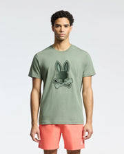 A man models against a plain backdrop in a Psycho Bunny MENS NEW CASTLE GRAPHIC TEE - B6U415F200 with a bunny and crossbones design, made of soft Pima cotton, paired effortlessly with coral shorts.