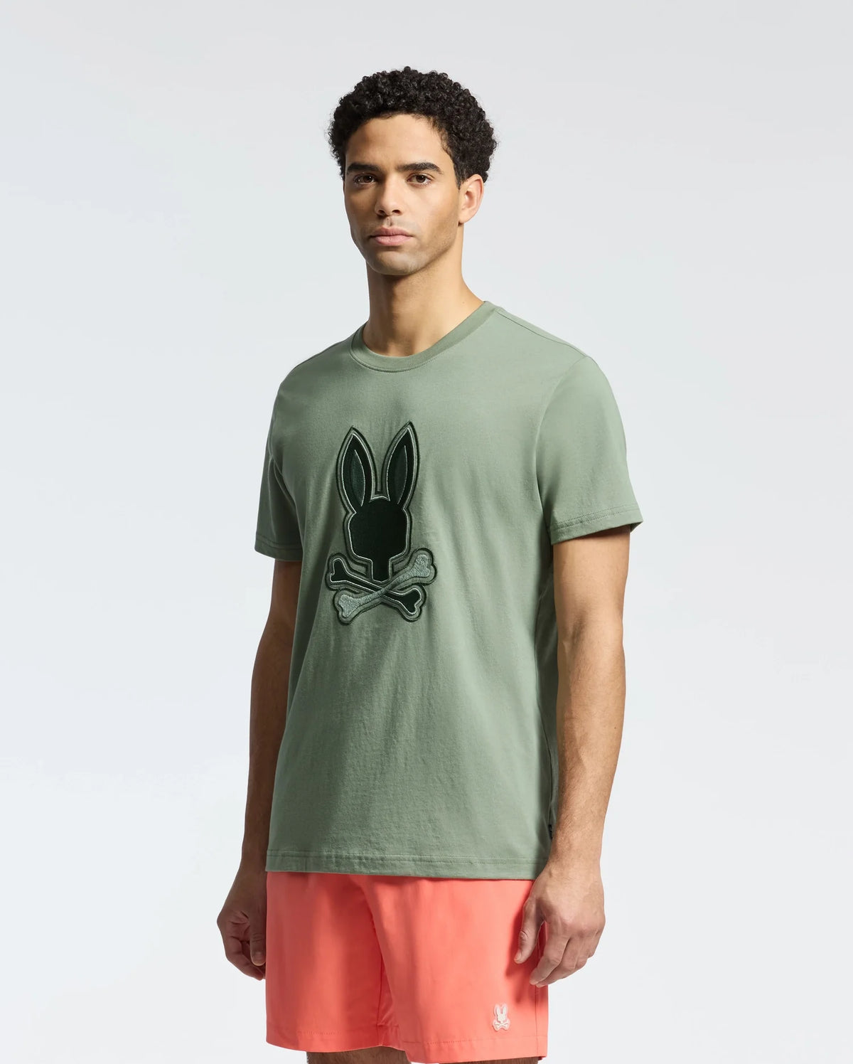 A man with short curly hair wears a sage green Psycho Bunny Men's New Castle Graphic Tee (B6U415F200) featuring an embroidered bunny and crossbones on the front, paired with coral shorts, against a plain white background.