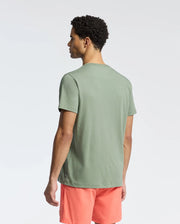 A man in a Psycho Bunny MENS NEW CASTLE GRAPHIC TEE - B6U415F200 and coral shorts stands with his back to the camera against a plain background.