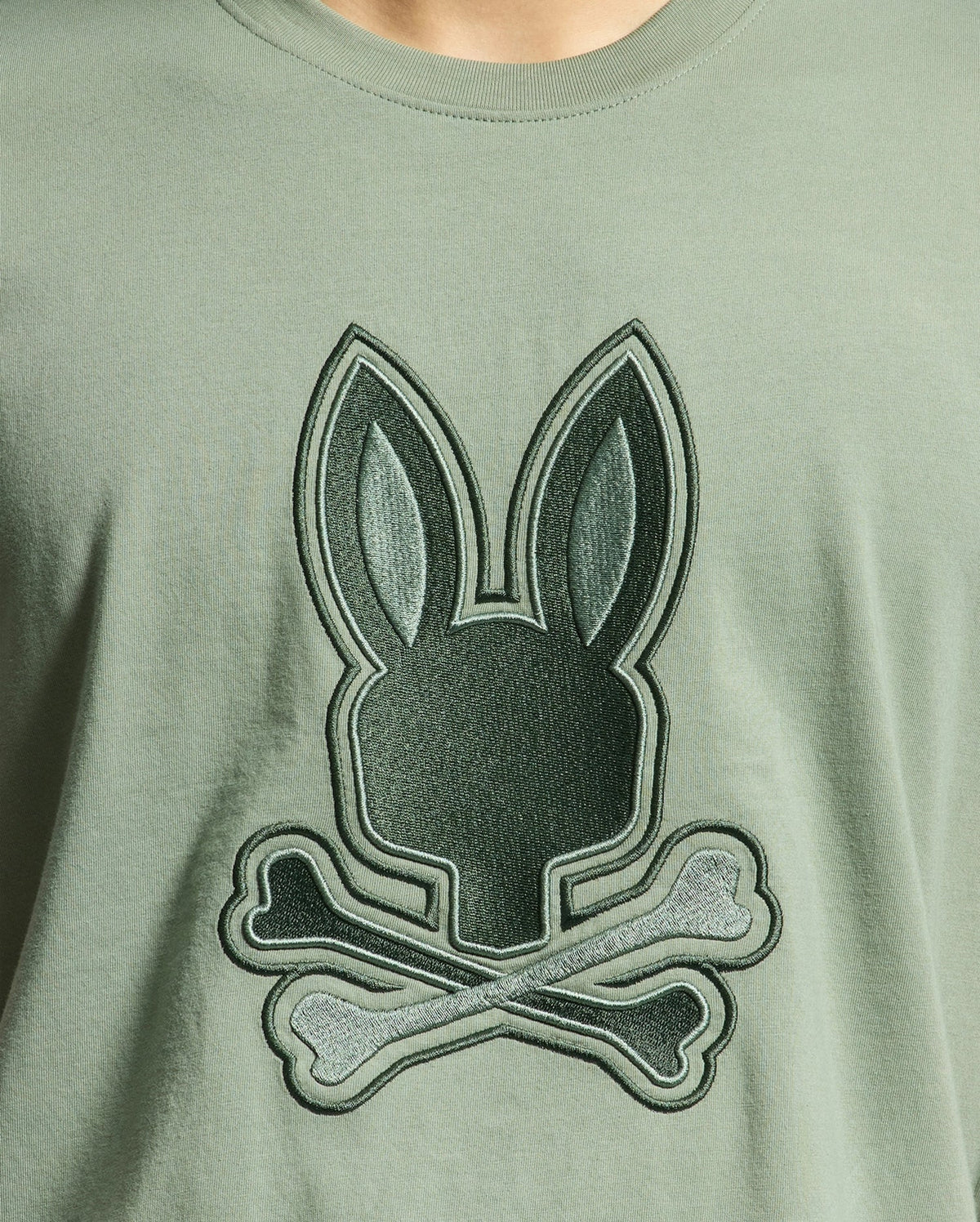 The Psycho Bunny MENS NEW CASTLE GRAPHIC TEE - B6U415F200 in green features a close-up of an embroidered bunny head with long ears above crossed bones, made from soft Pima cotton.