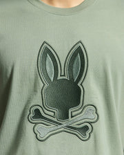 The Psycho Bunny MENS NEW CASTLE GRAPHIC TEE - B6U415F200 in green features a close-up of an embroidered bunny head with long ears above crossed bones, made from soft Pima cotton.