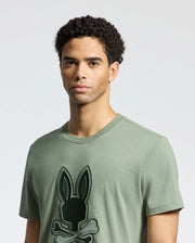 A person with curly hair is wearing a Psycho Bunny MENS NEW CASTLE GRAPHIC TEE in olive green made of soft Pima cotton, featuring an embroidered black bunny with crossbones on a plain, light-colored background.