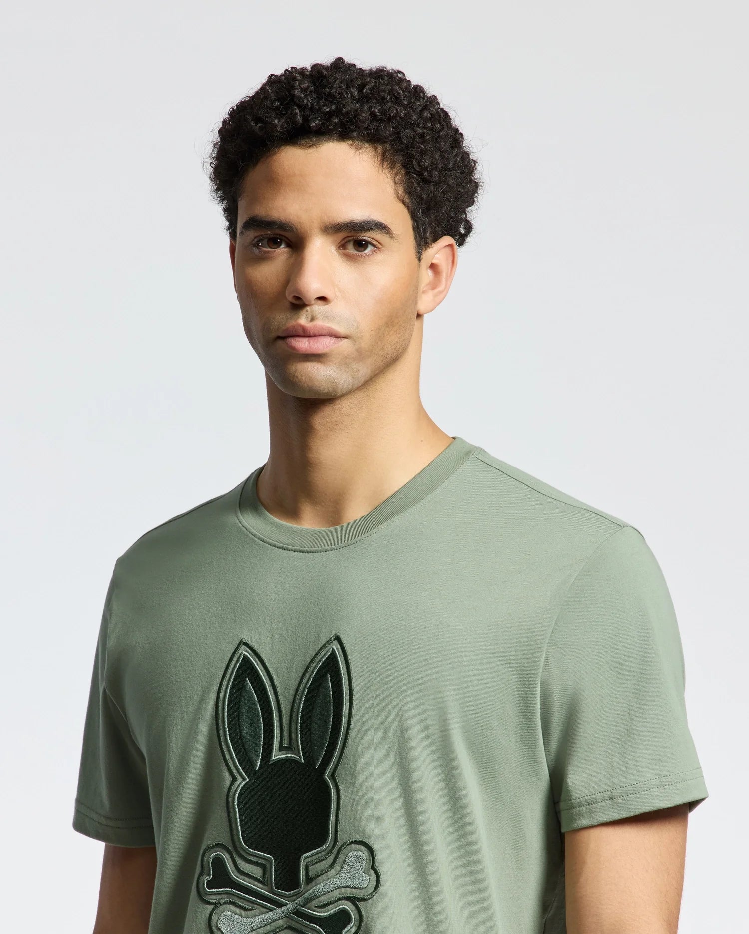 A person with curly hair is wearing a Psycho Bunny MENS NEW CASTLE GRAPHIC TEE in olive green made of soft Pima cotton, featuring an embroidered black bunny with crossbones on a plain, light-colored background.