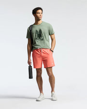 A person stands against a plain background wearing a Psycho Bunny MENS NEW CASTLE GRAPHIC TEE in green, coral shorts, and white sneakers. They hold a black water bottle and have a bag strap on their shoulder.