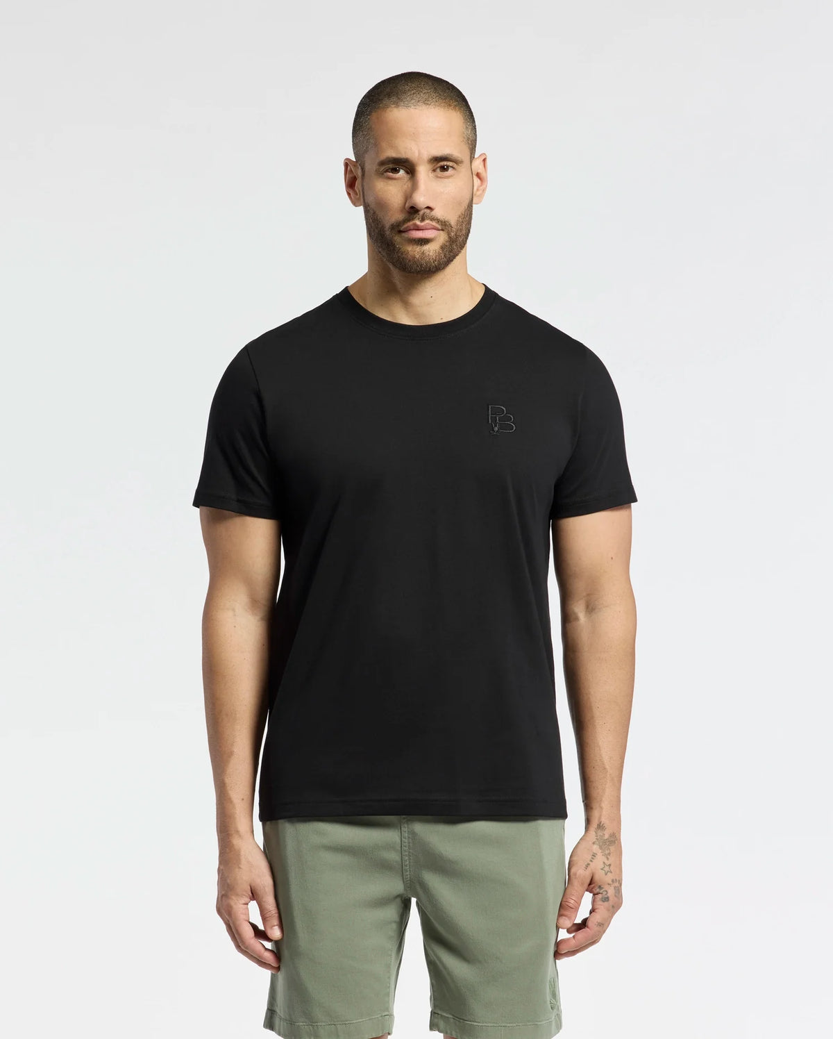 A man in a regular-fit Psycho Bunny MENS NICHOLSON PB CHEST GRAPHIC TEE (B6U416F200), made of Pima cotton, and green shorts stands against a plain white background. His arms rest by his sides, highlighting the shirt's smooth texture while maintaining a neutral expression.