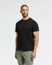 A man with short hair and a beard models a Psycho Bunny MENS NICHOLSON PB CHEST GRAPHIC TEE in black, teamed with green shorts. Embroidered details enhance his tattooed left forearm against a plain white background.