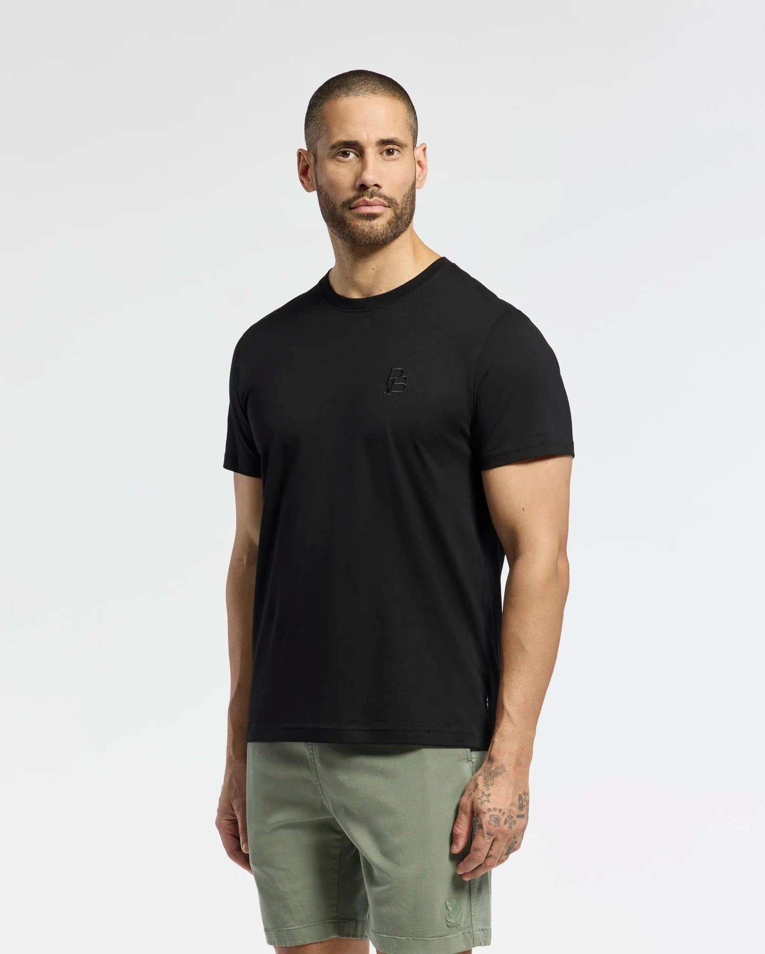 A man with short hair and a beard models a Psycho Bunny MENS NICHOLSON PB CHEST GRAPHIC TEE in black, teamed with green shorts. Embroidered details enhance his tattooed left forearm against a plain white background.