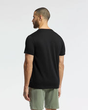A man with short hair stands facing away against a white background, wearing a plain black Psycho Bunny Pima cotton T-shirt and light green shorts, both in a regular fit.