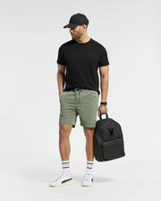 A man in a black cap and green shorts, wearing a Psycho Bunny Mens Nicholson PB Chest Graphic Tee, stands with a black backpack. He sports white sneakers with black stripes and a tattoo on his left arm, featuring embroidered details. He gazes sideways against a plain background.