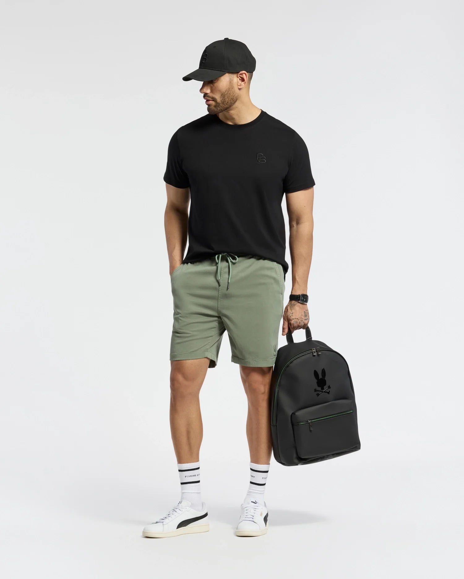 A man in a black cap and green shorts, wearing a Psycho Bunny Mens Nicholson PB Chest Graphic Tee, stands with a black backpack. He sports white sneakers with black stripes and a tattoo on his left arm, featuring embroidered details. He gazes sideways against a plain background.