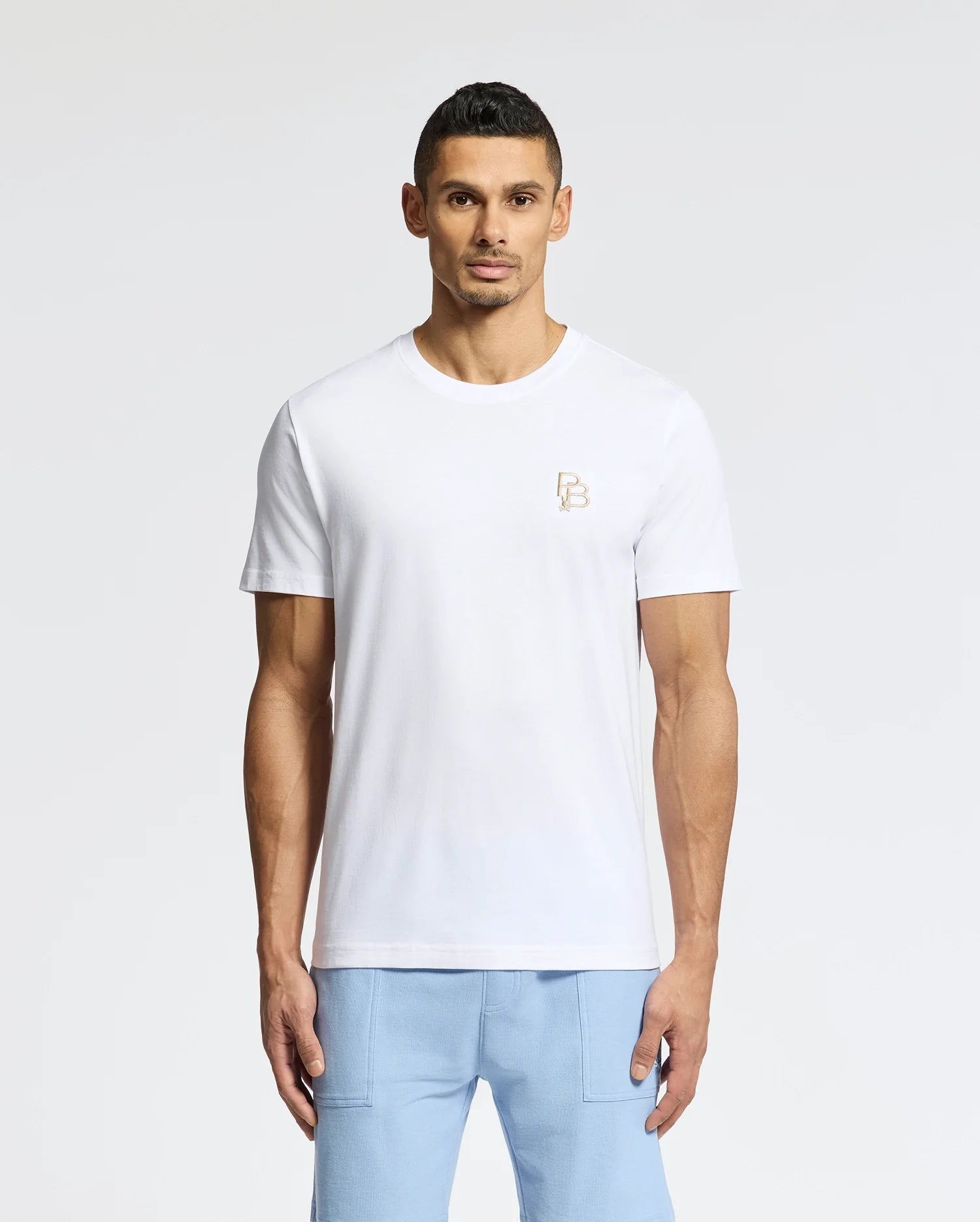A man is wearing the Psycho Bunny MENS NICHOLSON PB CHEST GRAPHIC TEE - B6U416F200, paired with light blue pants. The background is a plain, light color.