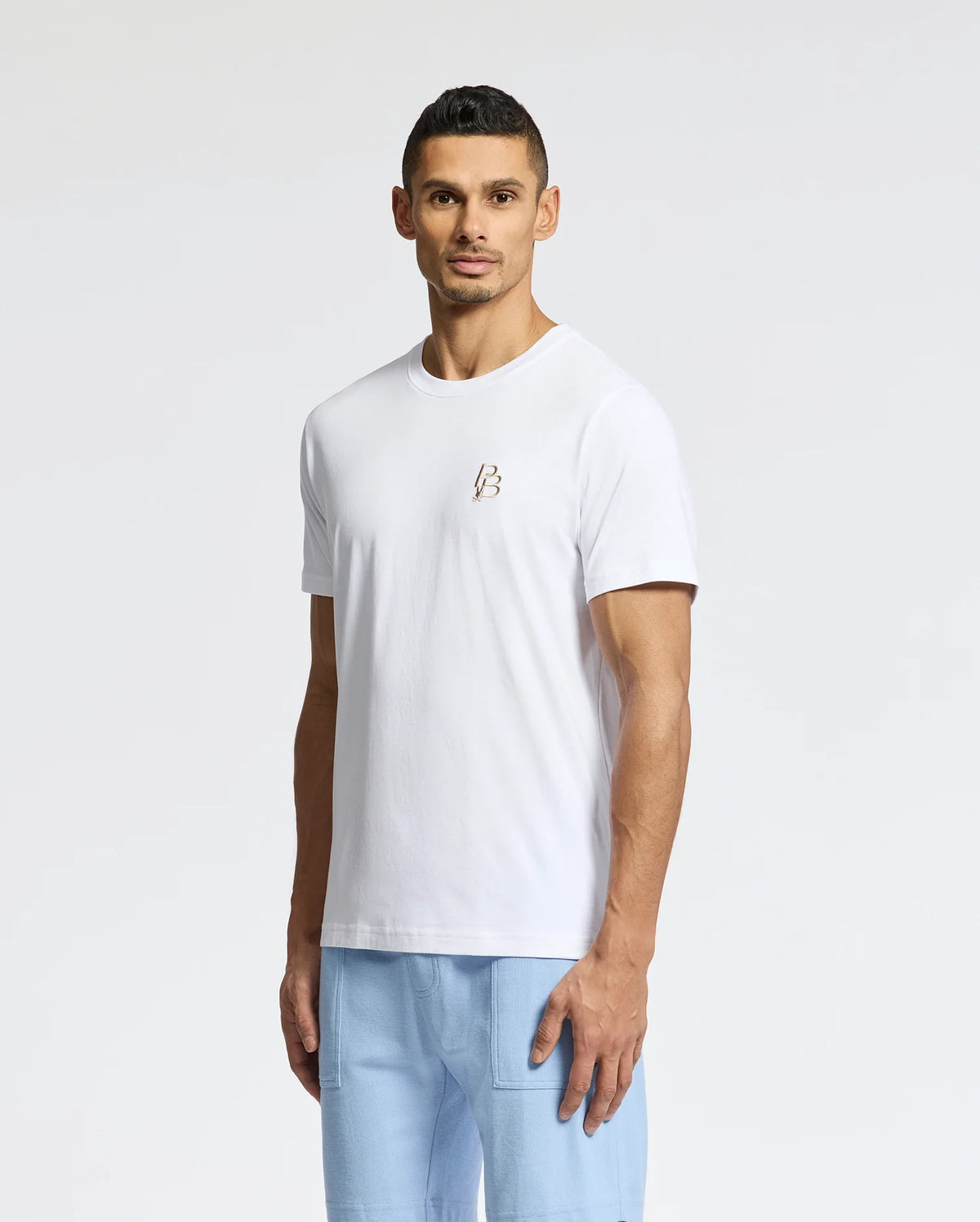 A man wearing a Psycho Bunny MENS NICHOLSON PB CHEST GRAPHIC TEE in white and light blue pants stands against a plain white background.