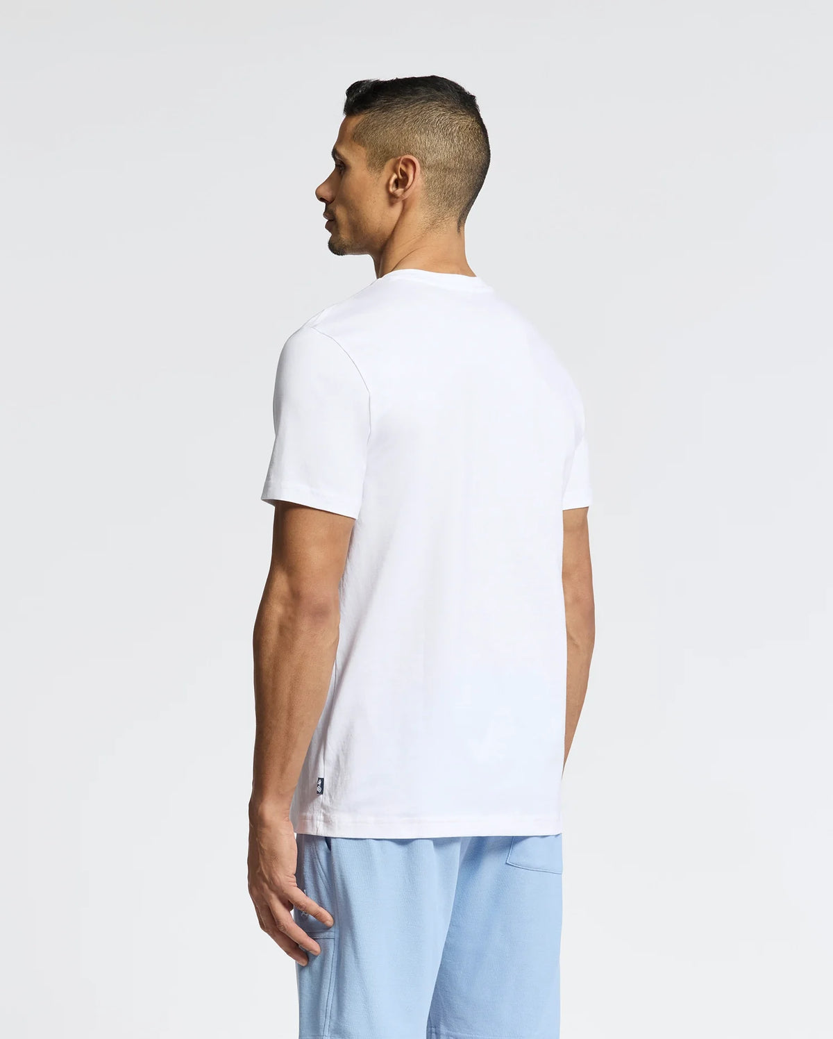 A person with short dark hair stands sideways, wearing the Psycho Bunny MENS NICHOLSON PB CHEST GRAPHIC TEE and light blue shorts, subtly facing away against a plain background.