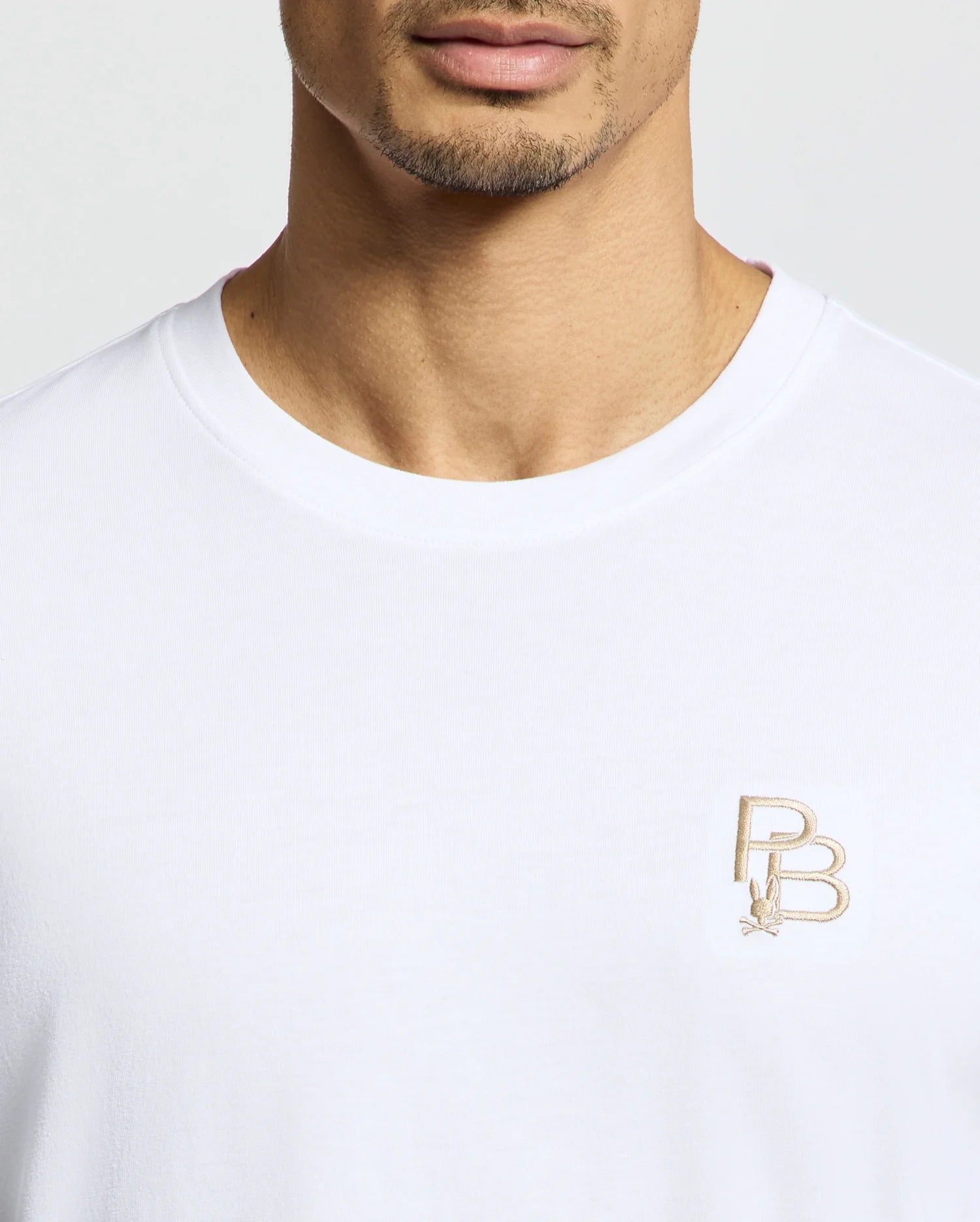 A man wears the Psycho Bunny MENS NICHOLSON PB CHEST GRAPHIC TEE - B6U416F200, a white Pima cotton tee with "PB" and a small embroidered leaf on the left chest. The image highlights his upper torso without any background details.