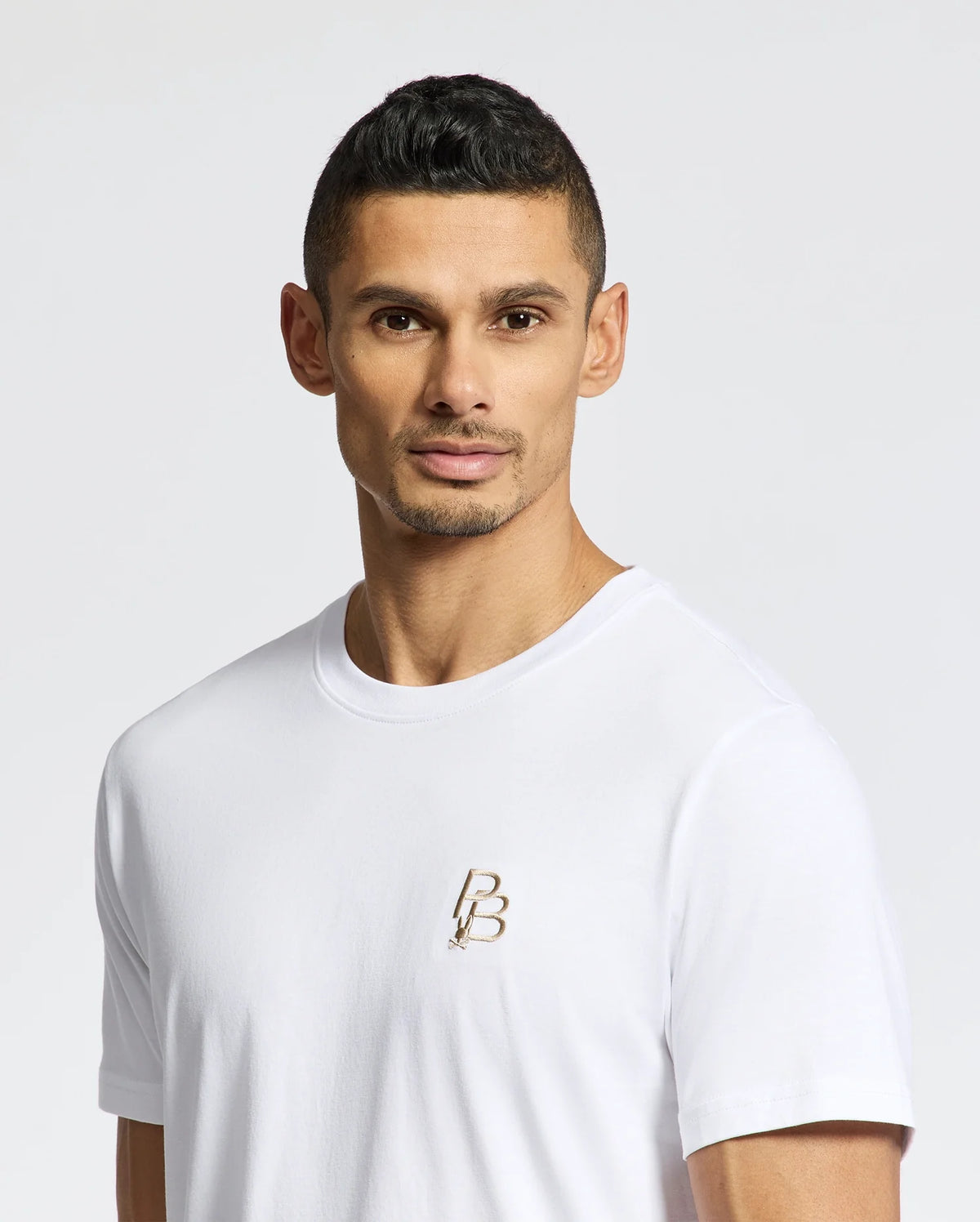 A person with short dark hair wears a Psycho Bunny's MEN'S NICHOLSON PB CHEST GRAPHIC TEE against a plain white background, looking directly at the camera.