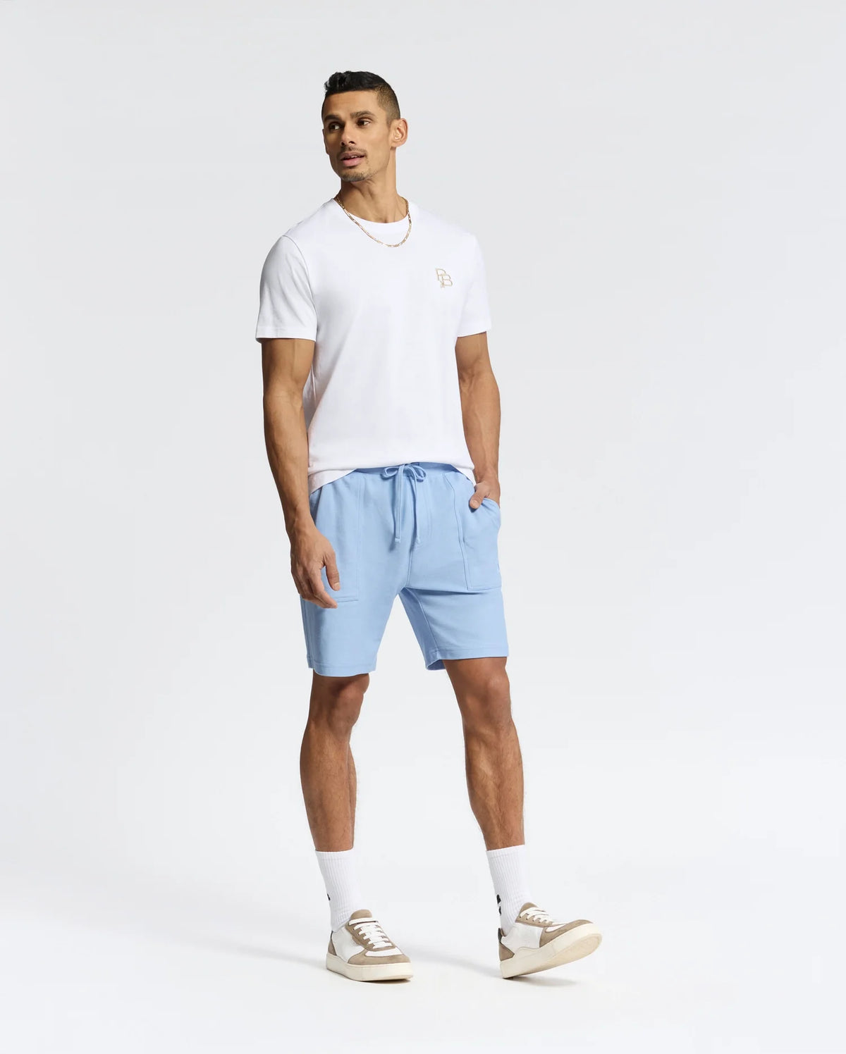 A man in a Psycho Bunny MENS NICHOLSON PB CHEST GRAPHIC TEE, light blue shorts, and white sneakers with beige accents stands against a plain white background. The Pima cotton shirt features an embroidered logo as he looks to the side with one hand in his pocket.
