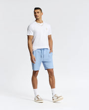 A man in a Psycho Bunny MENS NICHOLSON PB CHEST GRAPHIC TEE, light blue shorts, and white sneakers with beige accents stands against a plain white background. The Pima cotton shirt features an embroidered logo as he looks to the side with one hand in his pocket.