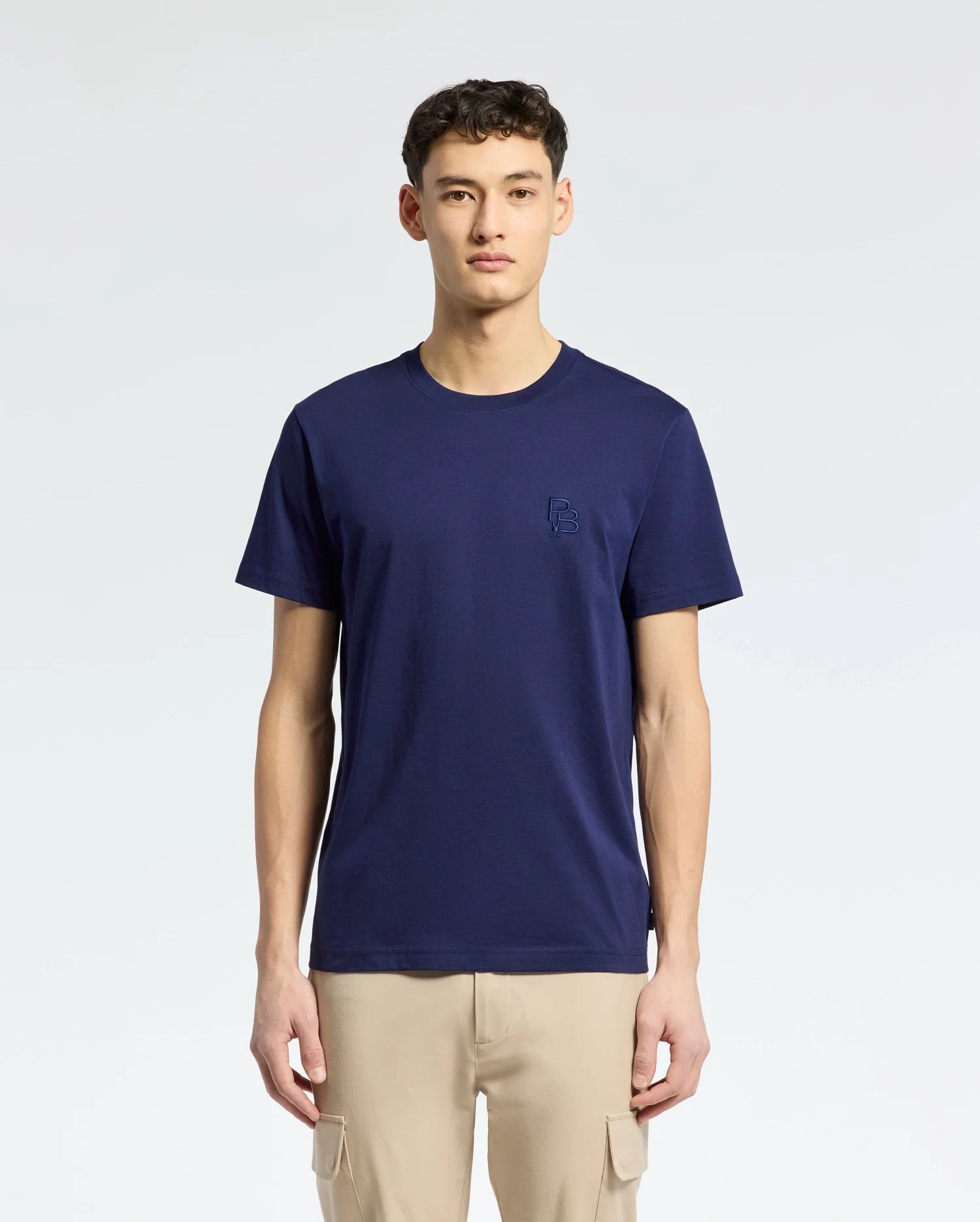A person with short dark hair wears a navy blue Psycho Bunny MENS NICHOLSON PB CHEST GRAPHIC TEE - B6U416F200, crafted from Pima cotton and featuring embroidered details with a small logo on the left chest. They're in beige cargo pants against a plain light background, looking directly at the camera.