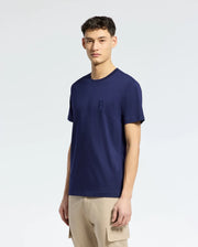 A man wears a Psycho Bunny MENS NICHOLSON PB CHEST GRAPHIC TEE (B6U416F200), a plain navy blue Pima cotton tee with an embroidered logo, paired with beige cargo pants against a simple light gradient backdrop.