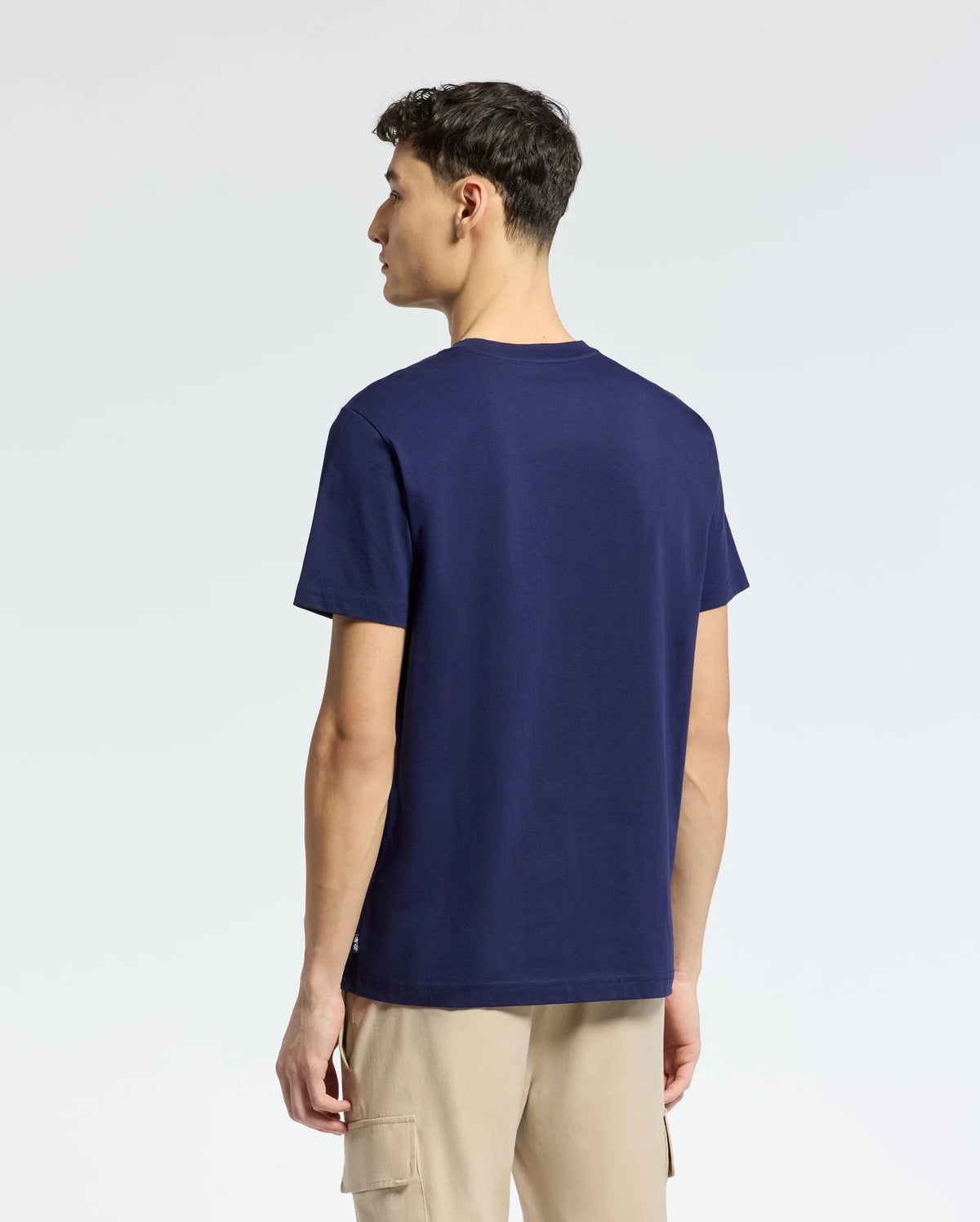 A person in a Psycho Bunny MENS NICHOLSON PB CHEST GRAPHIC TEE - B6U416F200, navy blue with embroidered details, and beige regular-fit pants is standing and looking left against a light, plain background.
