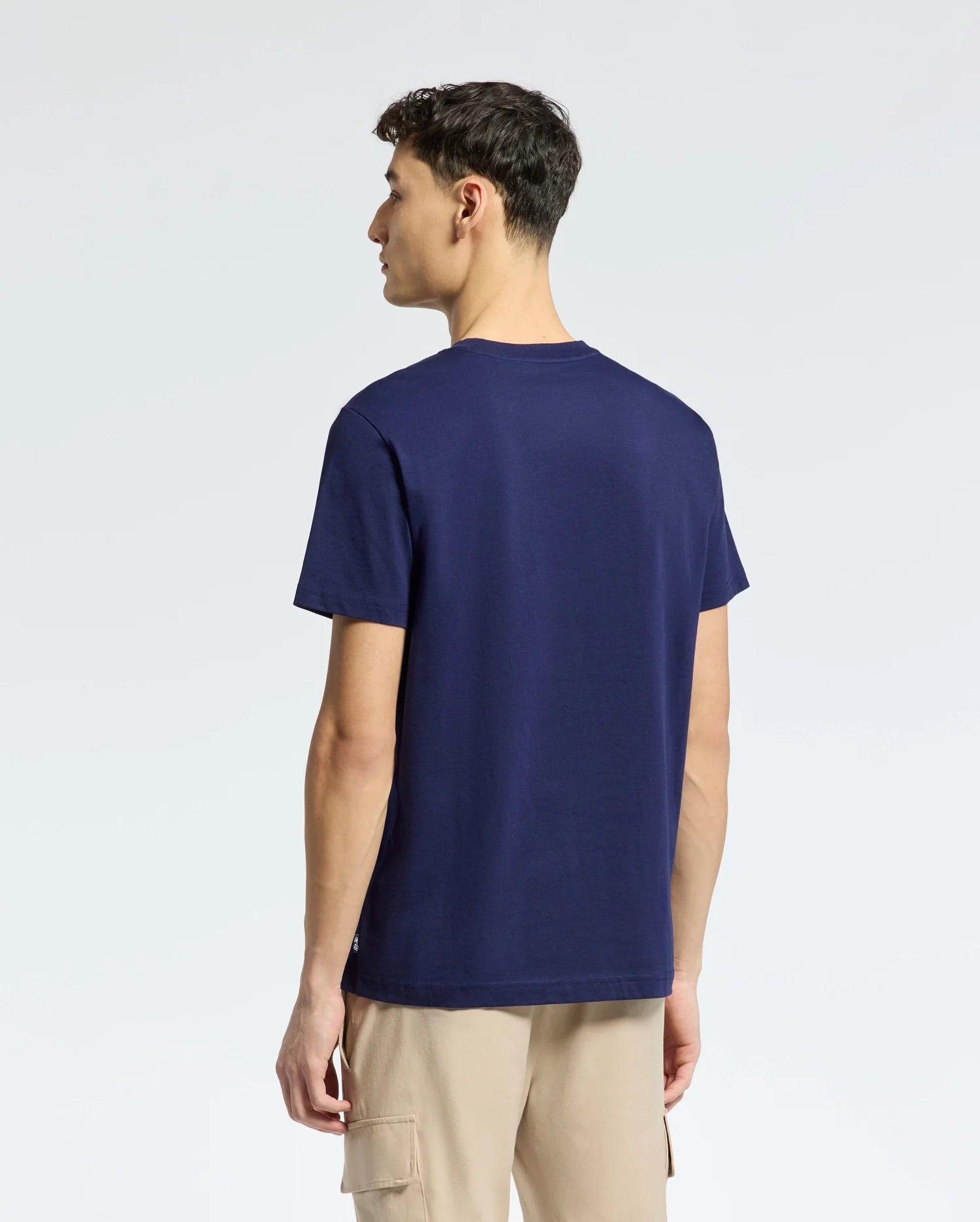 A person in a Psycho Bunny MENS NICHOLSON PB CHEST GRAPHIC TEE - B6U416F200, navy blue with embroidered details, and beige regular-fit pants is standing and looking left against a light, plain background.