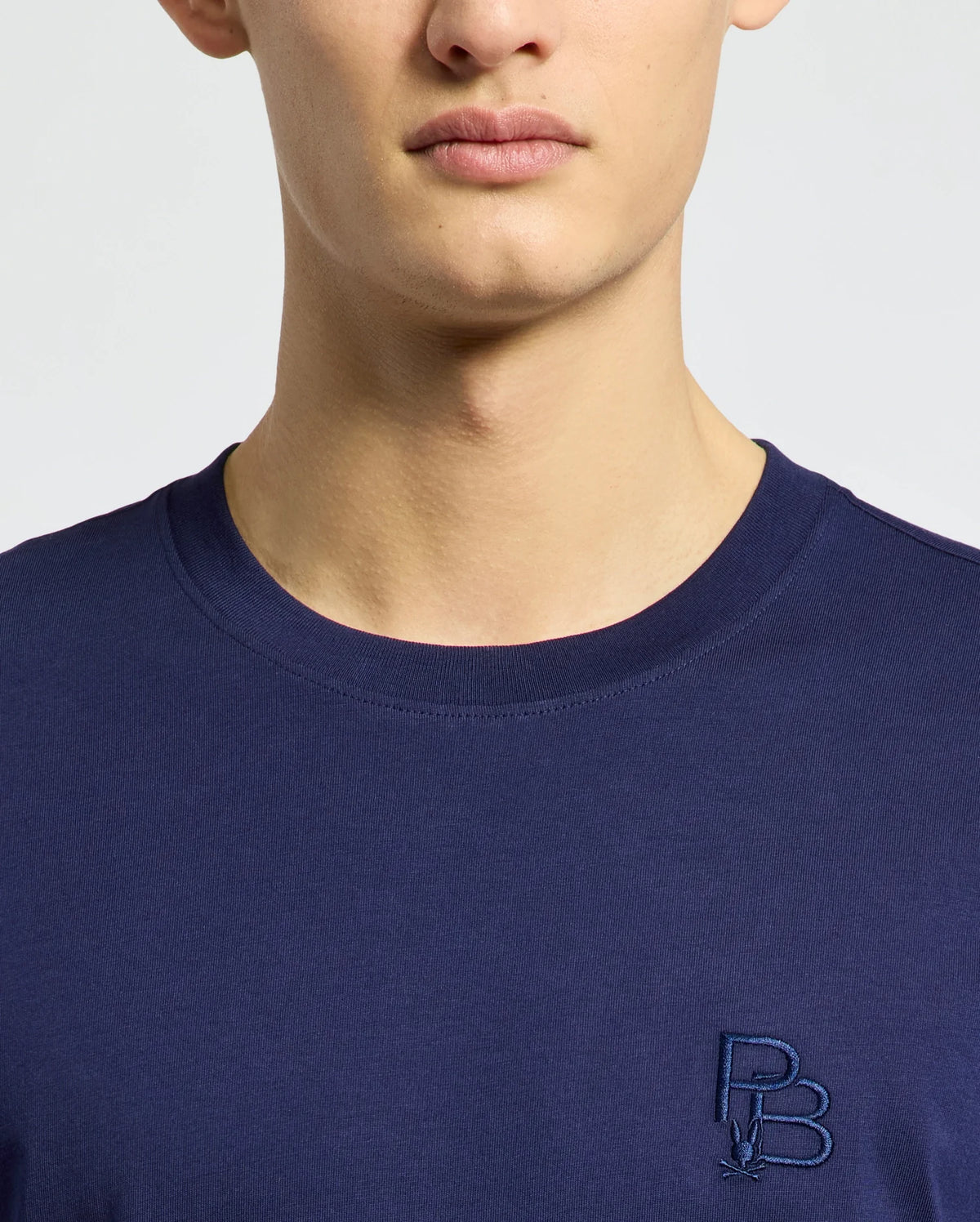 A person is wearing the Psycho Bunny MENS NICHOLSON PB CHEST GRAPHIC TEE - B6U416F200, a navy blue regular fit Pima cotton t-shirt with "PB" initials and a small chest graphic featuring embroidered details, standing against a plain light background.