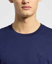 A person is wearing the Psycho Bunny MENS NICHOLSON PB CHEST GRAPHIC TEE - B6U416F200, a navy blue regular fit Pima cotton t-shirt with "PB" initials and a small chest graphic featuring embroidered details, standing against a plain light background.